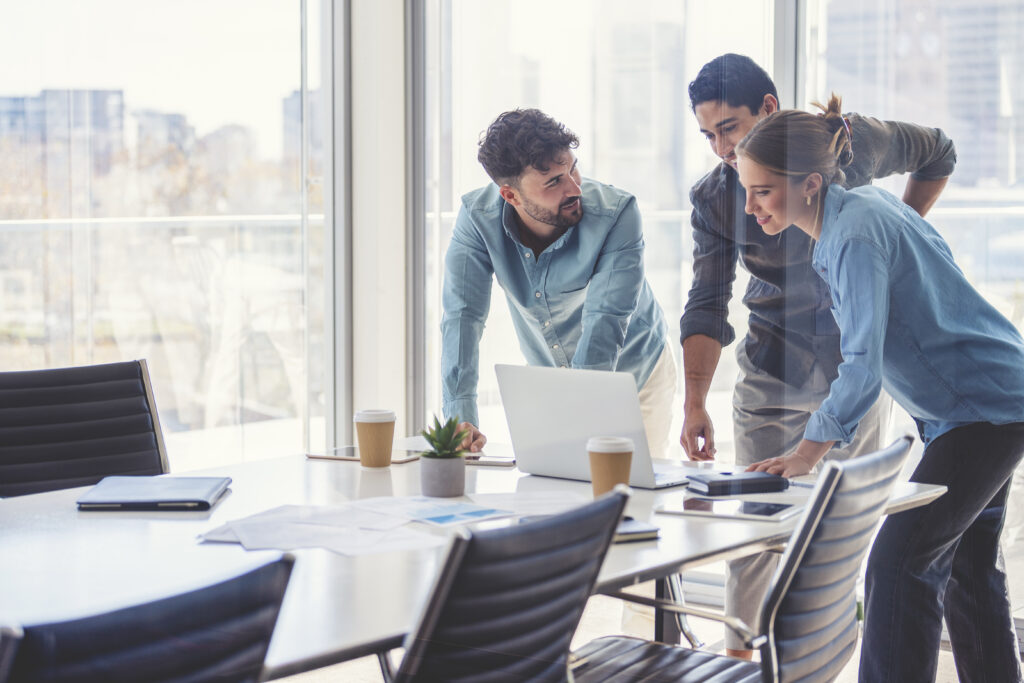 Business team working on a laptop computer. Three people are wearing  casual clothing. They are standing in a board room. Multi ethnic group with Caucasian and Latino men and women. They are all happy and smiling